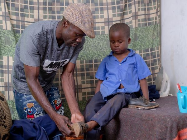 Alt-text: Derrick, a father in Zambia, helps his toddler son Abraham put on his shoes