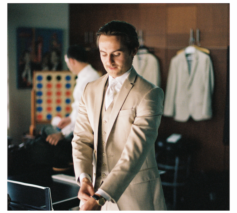 Groom adjusting cufflinks while wearing a light tan suit with a white tie in a dressing room
