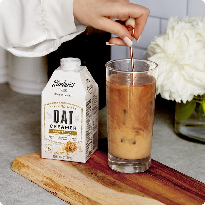 A person stirring Elmhurst Brown Sugar Oat Creamer into a tall glass of iced coffee with a gold spoon.