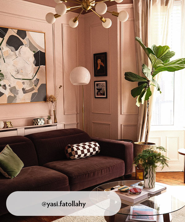 A softly lit living room with blush-pink panelled walls, a deep plum sofa, abstract art, and leafy plants. Brass lighting and cosy textures give the space a gentle elegance, blending modern style with comforting warmth.