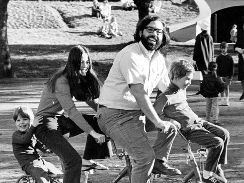 Four figures sit on a four person bike, a man and woman in the middle, and two young children on the outsides. They smile and ride through a park.