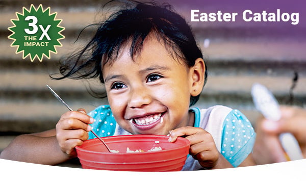 A little girl smiles while eating rice from a big red bowl.