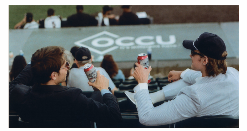 Two men sit in stadium seats watching a baseball game. One wears a navy suit, the other a light blue seersucker suit and cap. They raise canned drinks in a toast, enjoying the sunny day.