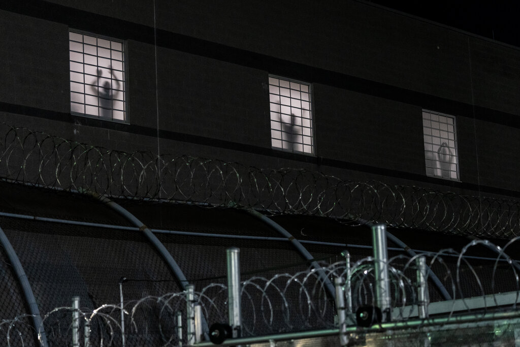 A dark building’s three lit windows show silhouettes of people behind grates. Razor wire is in the foreground.
