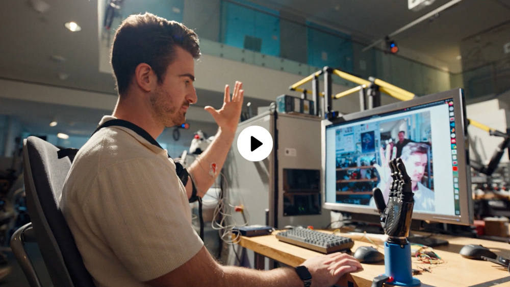 A researcher holds their left hand up, mimicking the robotic hand on table, while looking at a computer screen that shows themselves on camera