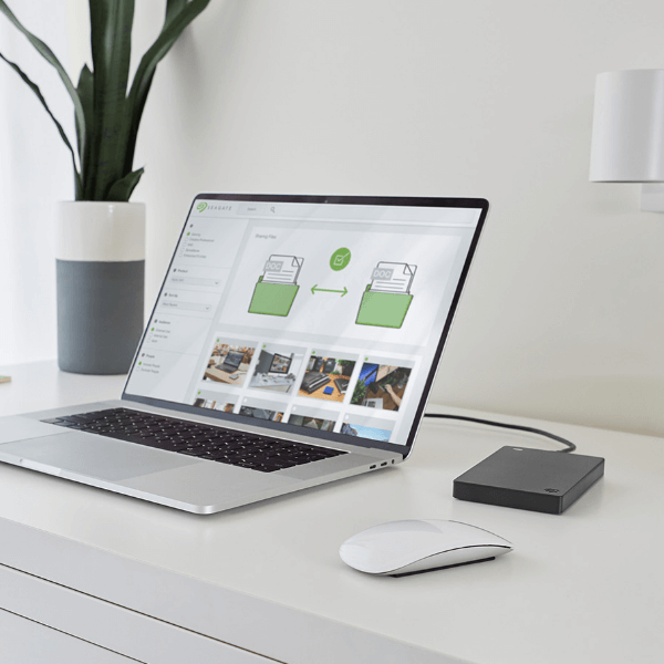 A sleek laptop on a modern white desk. Next to it are a white mouse and a black external hard drive.