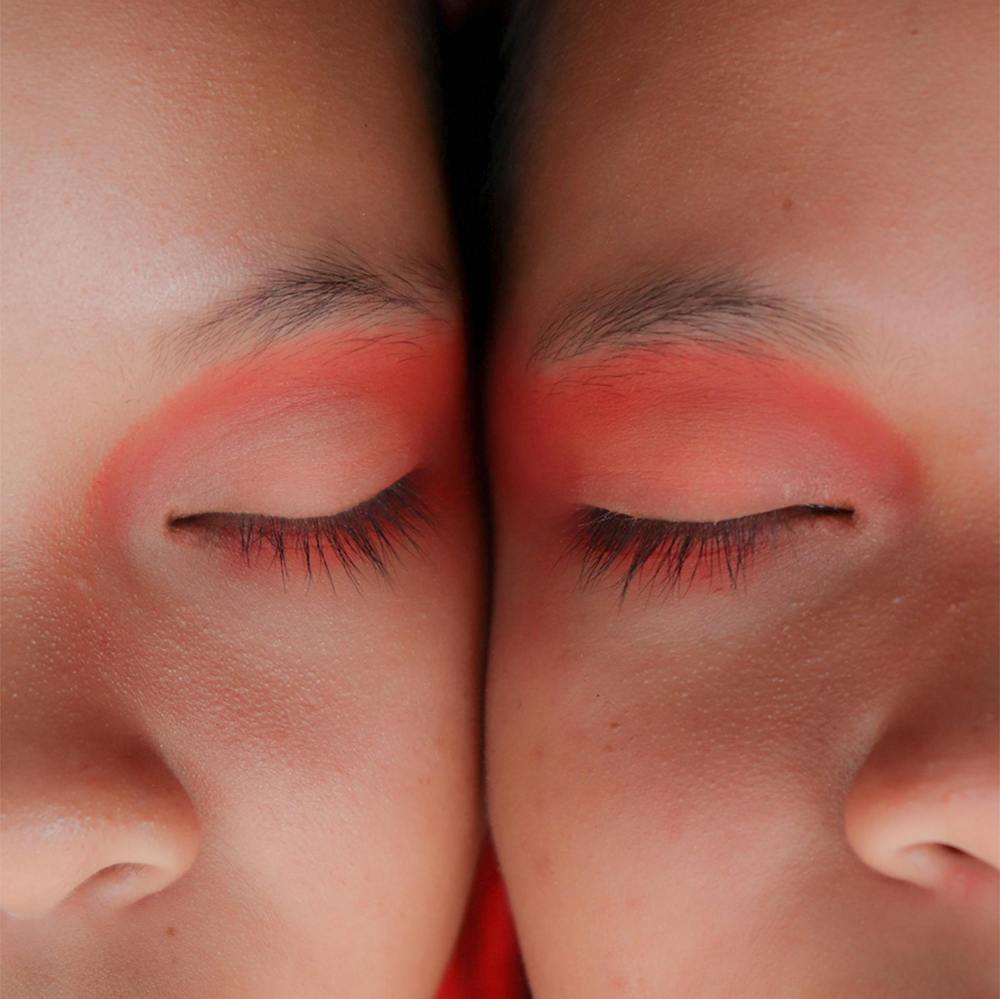 A close-up on two people with very similar faces with their cheeks pressed together, their eyes closed. They have pinkish-orange eye-shadow on. 