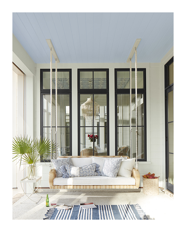 A white porch with window trim painted black, a white floor, blue and white area rug, and a hanging swing.