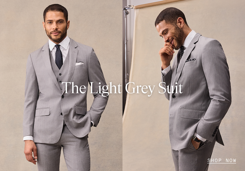  Two views of a man wearing a light grey three-piece suit — a front-facing shot styled with a navy dotted tie and white pocket square, and a three-quarter view with a dotted tie and pocket square. The Light Grey Suit. SHOP NOW