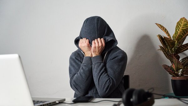 A person covering his face with his hands and a hoodie at an office desk.