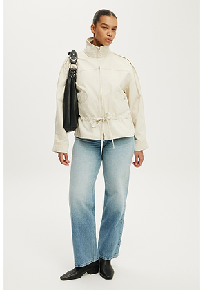 Model wearing cream zip-up jacket with drawstring waist and light blue jeans, holding black shoulder bag