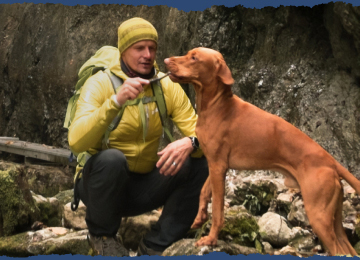 A hiker in a yellow jacket and beanie is feeding his excited dog a treat. They’re in a shaded area with moss-covered rocks.