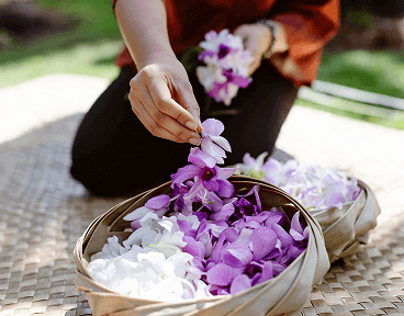 A person carefully crafting leis, placing vibrant purple and white flowers into a basket.