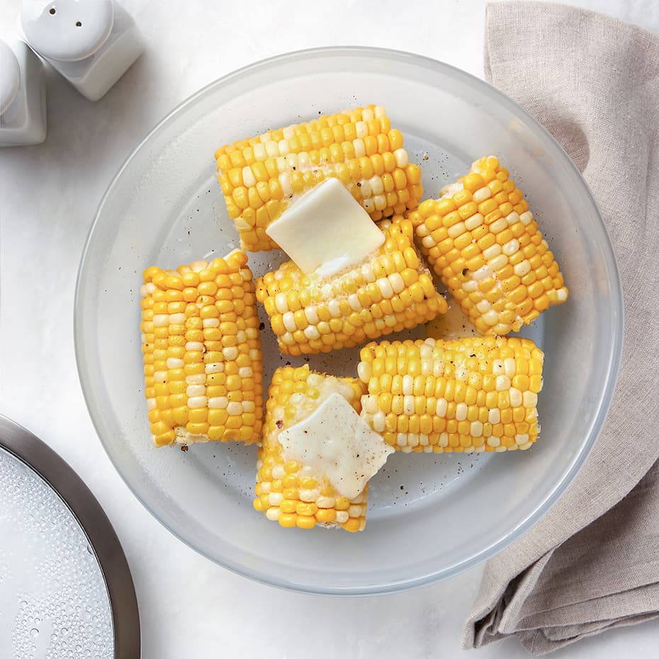 Overhead view of a round Anyday glass dish filled with cooked corn on the cob topped with melting pats of butter, with a beige napkin and salt and pepper shakers in the background.