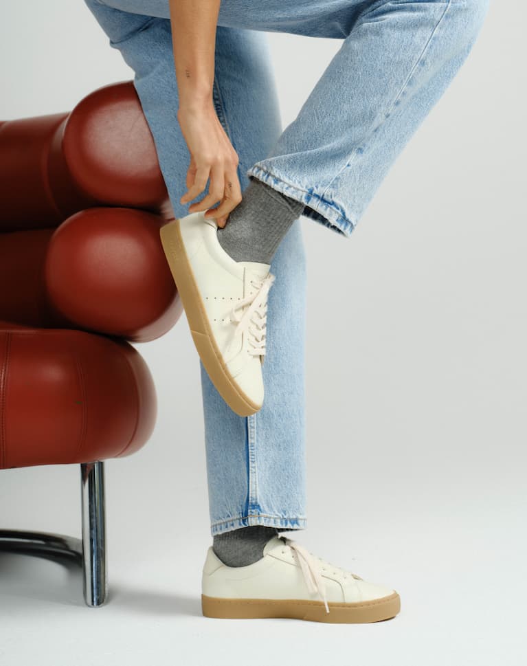 Girl standing next to red leather chair wearing light wash jeans, grey socks and white diamond cup sneakers