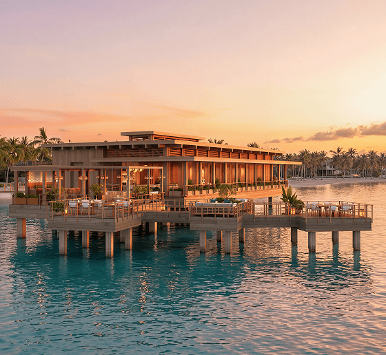 Overwater restaurant at sunset, with wooden decks, dining tables, and palm-lined shoreline in the background.