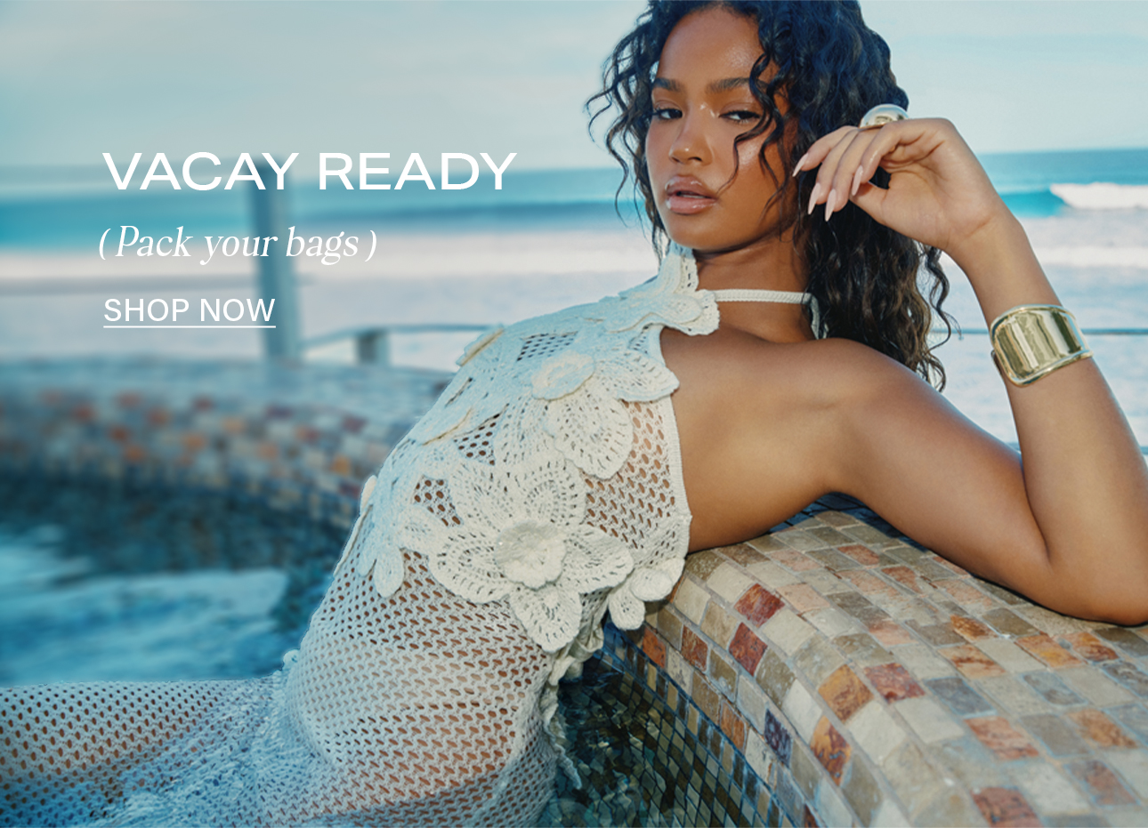Vacay Ready: Model in white crochet floral halter dress & gold accessories by the ocean. Shop trendy summer resort wear.