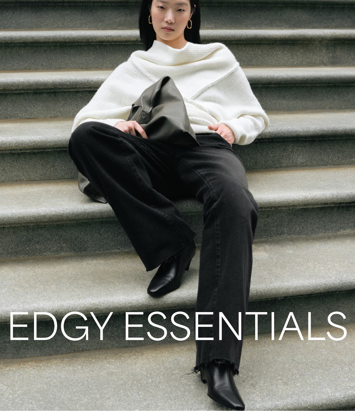 Xun sits on stone steps wearing The Palmer Long Sleeve Top, black wide-leg pants, pointed black boots, and holding a slouchy bag with the text “Edgy Essentials.”