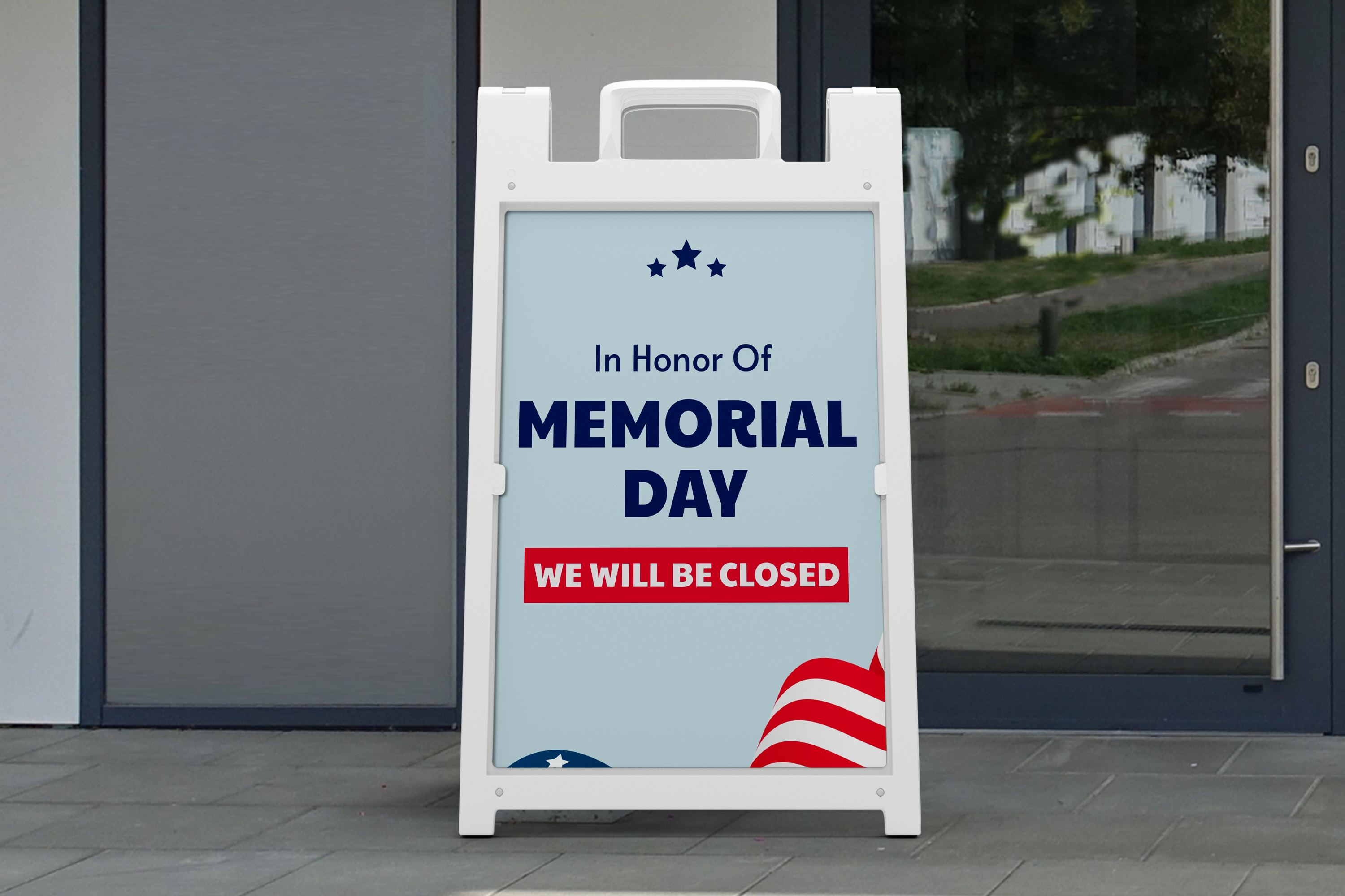  white A-frame  sign outside a building displays a Memorial Day closure notice with navy text reading “In Honor Of Memorial Day” and a red banner that says “WE WILL BE CLOSED,” accented with stars and flag-style graphics.