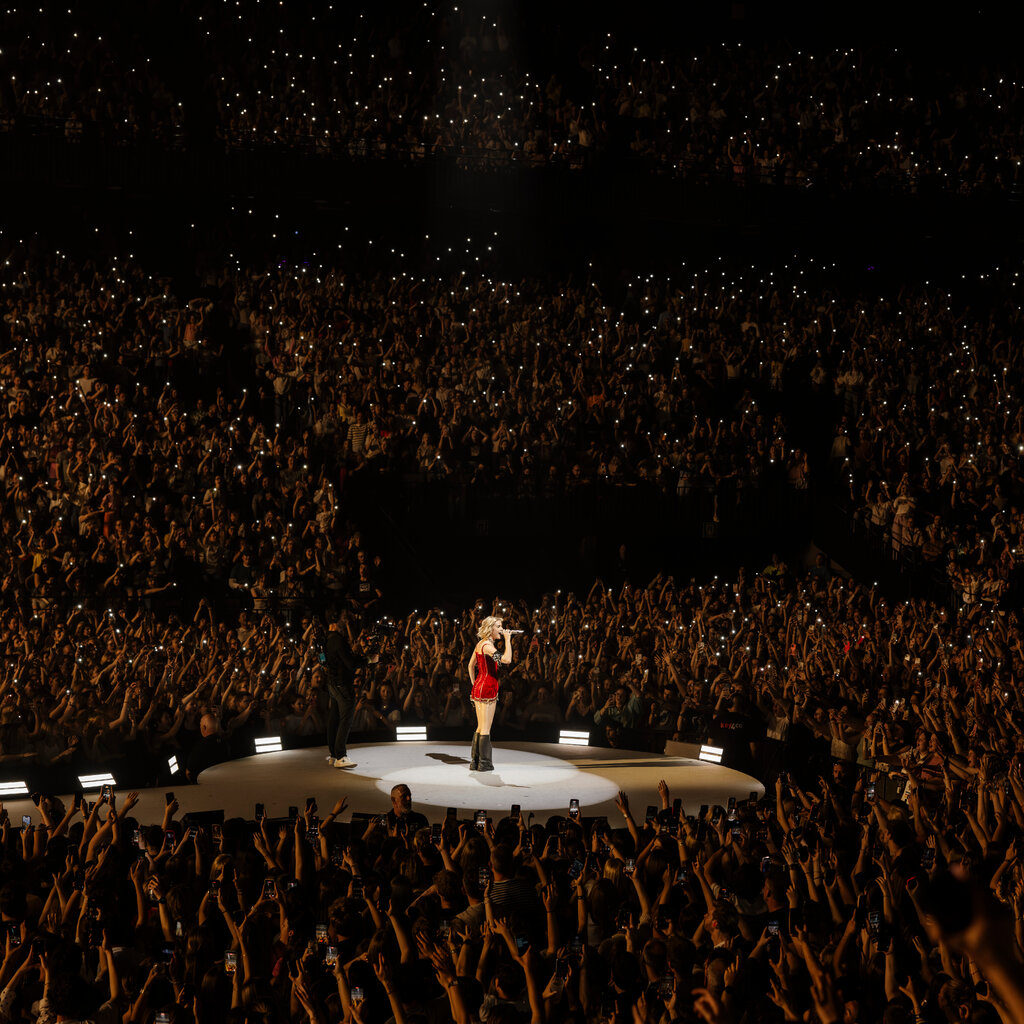 A woman in a red dress and black boots stands on a stage in the middle of a crowd.