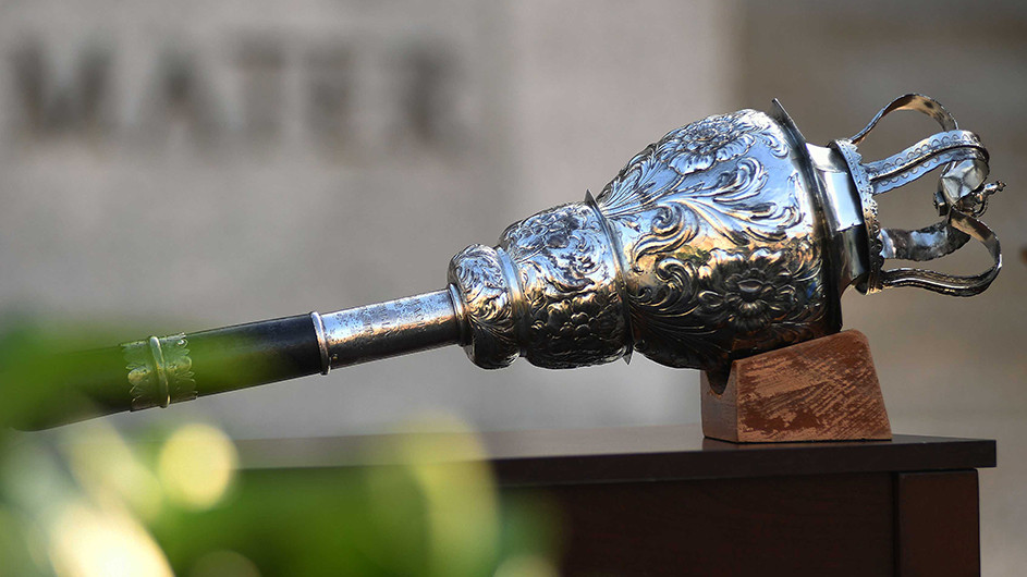 A roughly two-foot long wooden and silver-plated mace, adorned with acanthus leaves and topped with a crown, propped on a table in front of the statue of Alma Mater at Columbia University’s Commencement.