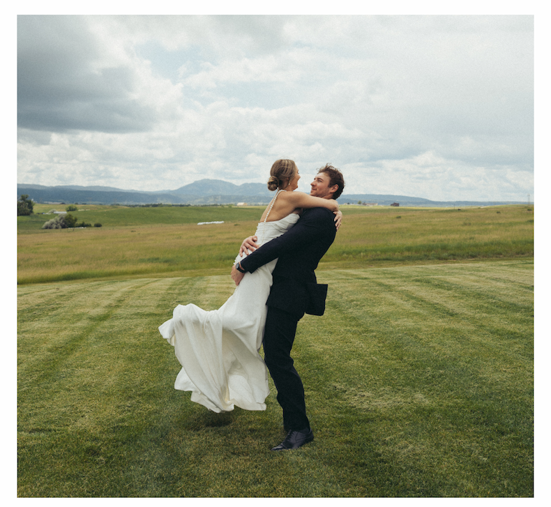 Groom lifting bride in a field with mountains in the background.