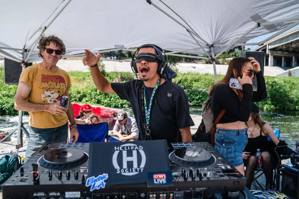 DJ Brawl wears headphones and holds his right arm up in the air as he performs at a DJ controller beneath a canopy, while others watch and relax nearby.