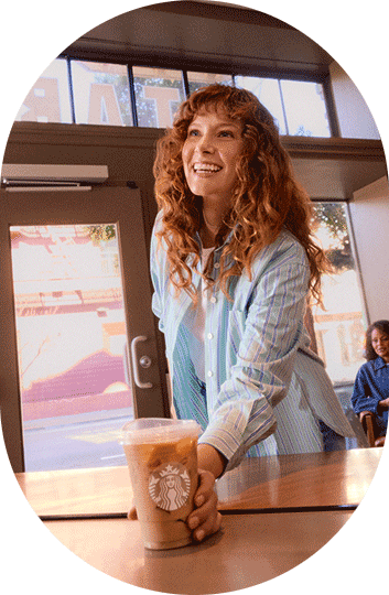 A smiling woman with long red hair picks up her iced coffee drink from the counter in a Starbucks cafe.