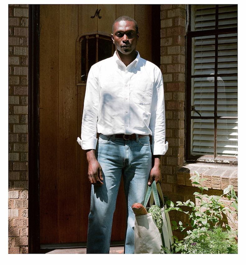 Man wearing a white oxford shirt and jeans, holding a tote bag with groceries including a baguette and greens.