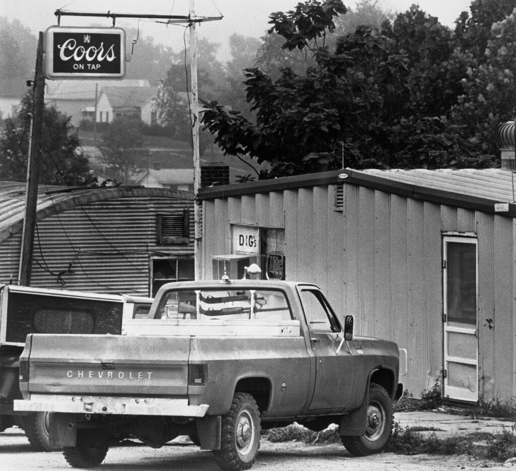 In a black-and-white image, a Chevrolet pickup truck is parked in front of a structure on a tree-lined street.