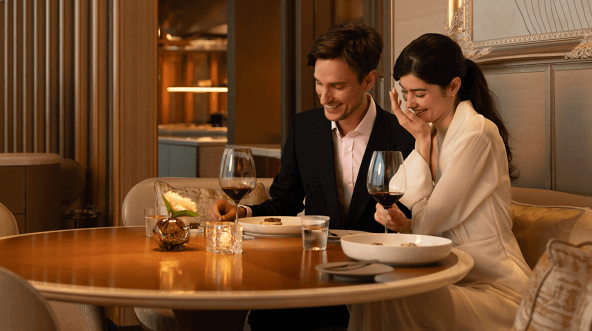 A couple sits in an elegant dining room, laughing over a meal and glasses of red wine.