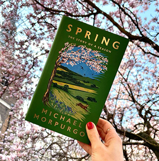 A green book is being held up against the sky, where the leaves of a cherry blossom tree can be seen in the background