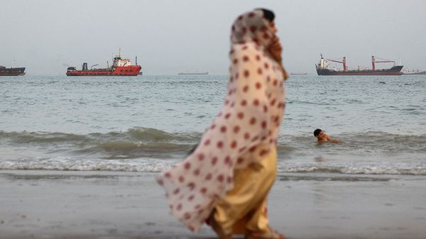 A woman walks on the beach while a boy swims, with vessels in the Strait of Hormuz visible near Bandar Abbas, Iran, May 22nd 2026