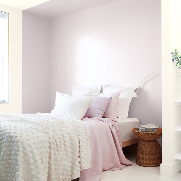 A lilac-painted bedroom with cozy linens, a wicker nightstand, and a variety of throw pillows.