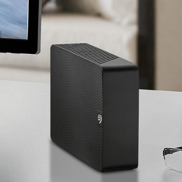 A Seagate Expansion Desktop sits on a desk beside a computer monitor and some glasses.