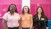 three students pose in front of pink background, with name tags that read, 
