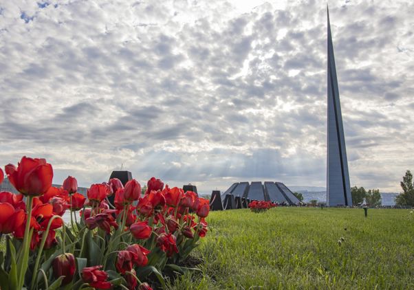 Red tulips bloom in the foreground of the Armenian Genocide Memorial Complex in Yerevan, where a tall spire rises beneath a cloud-filled sky with rays of sunlight breaking through.