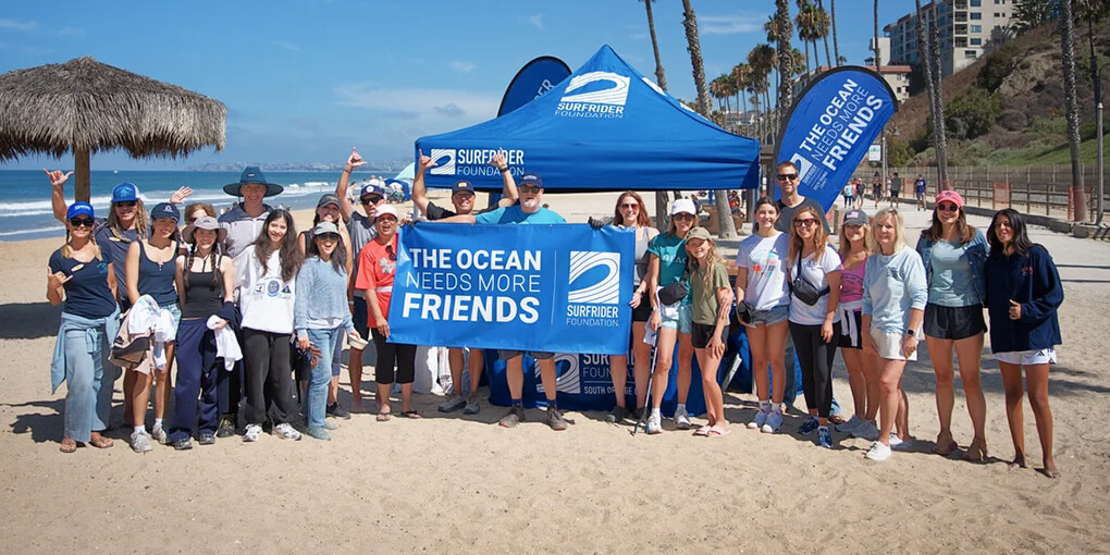 25 San Diego volunteers pose under a blue Surfrider tent with a sign that reads "The Ocean Needs More Friends."
