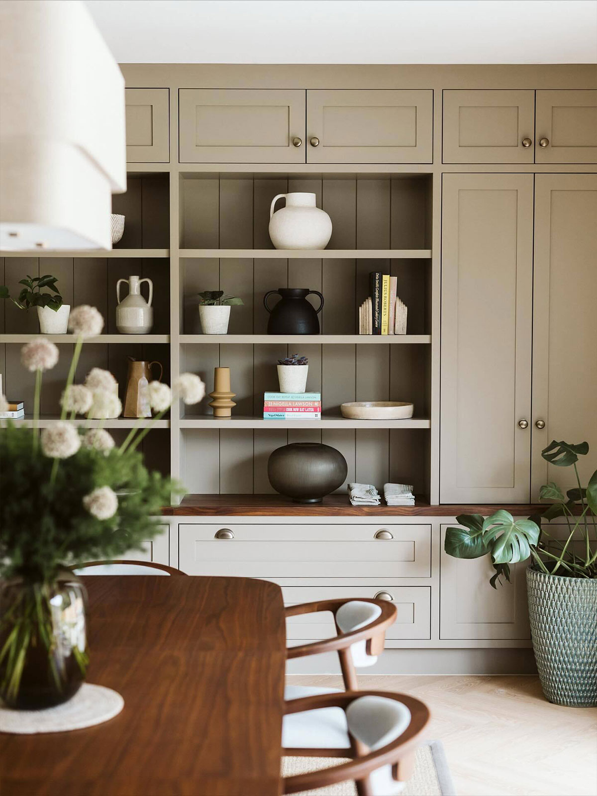 An elegant, beautifully organised dining space surrounded by built in shelves and cupboards, displaying curated treasures collected through the years. From a selection of ceramic jugs to few choice books held in place by minimalist bookends. Plants and flowers bring verdant softness to the quiet grey brown background, with white lightshades adding a touch of gentle brightness.