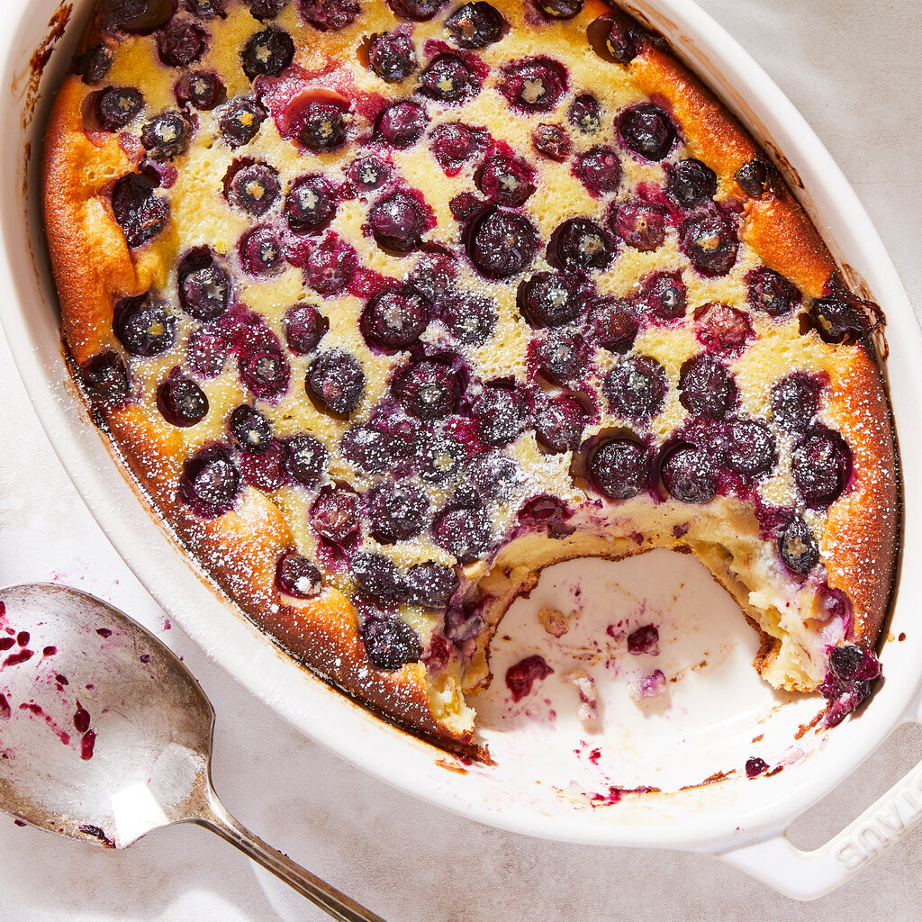 A golden-brown baked dessert topped with dark blueberries and powdered sugar, in a white oval dish.