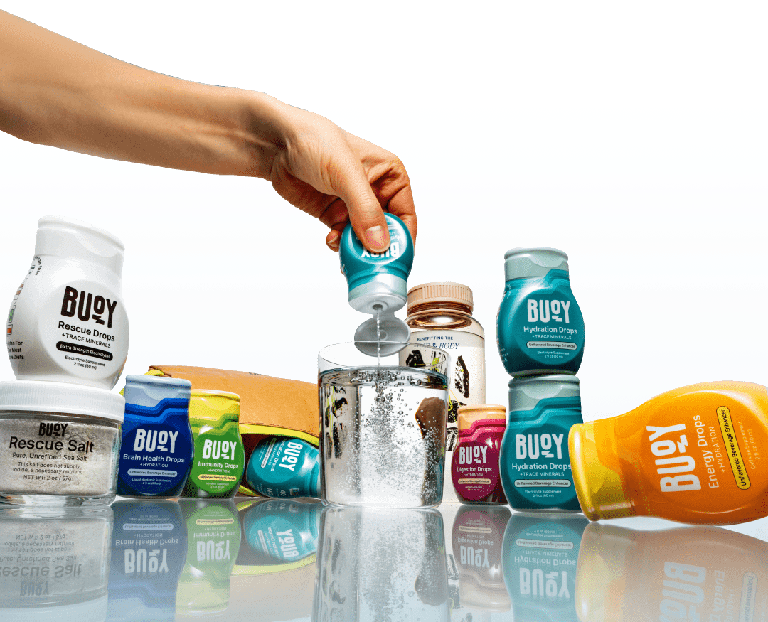 A hand squeezes Hydration Drops into a glass of water, surrounded by all Buoy products on a reflective surface.