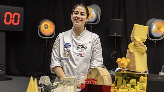  A female in a chef's coat stands behind a table filled with cheese.
