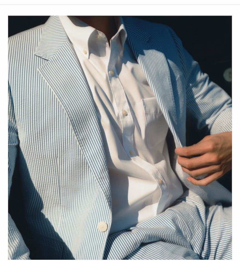 Close-up of a man seated in a pale blue and white seersucker suit. His white button-down shirt is neatly tucked in, and his hand lightly holds the lapel of the jacket.