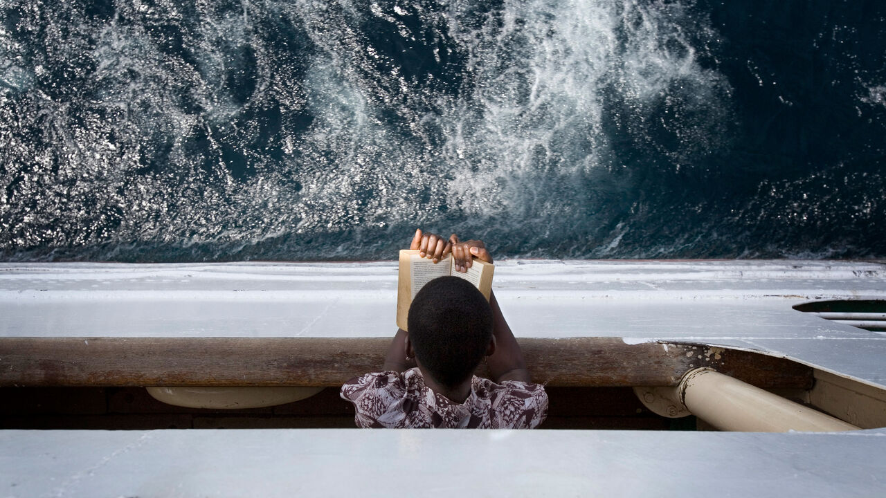 Woman reads a book leaning over a railing above the sea. 
