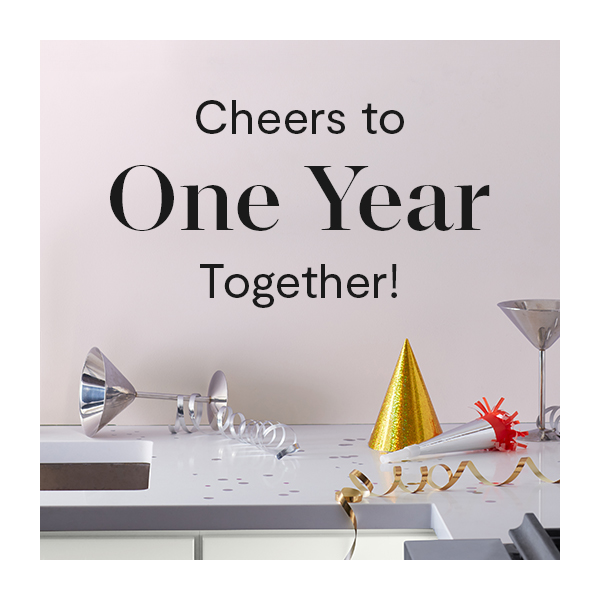 Cheers to One Year Together! Party supplies, including a gold metallic party hat and streamer, silvery party popper and two silver martini glasses, are strewn across a lilac countertop.