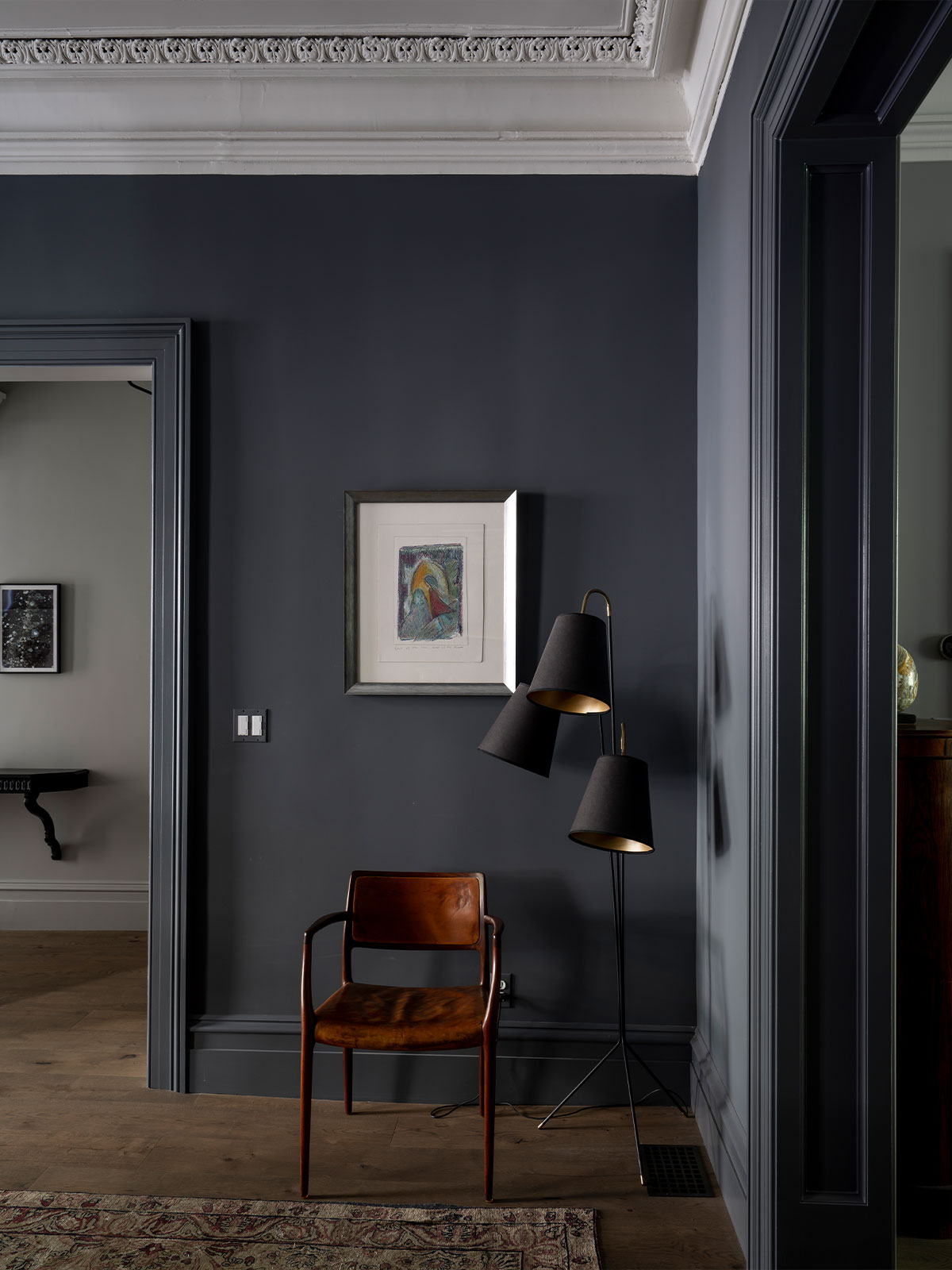 We find ourselves in a dramatic, moody, dark grey room with walls, woodwork and moulding all in the same shade, the perfect offset to the watercolour print on the wall. The lamp shades are dark grey but with a glowing pale gold interior visible on a couple of them. Beyond the archway, the walls are lighter grey, drawing us into that space to admire the small painting on the wall.