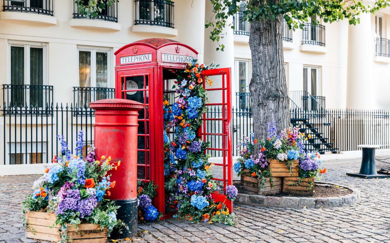 Classic red London telephone booth overflowing with vibrant flowers along a charming cobblestone street.