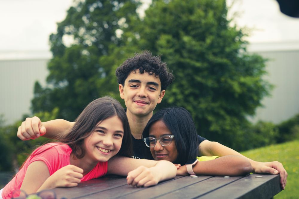 Three young people huddled round a picnic bench, smiling.