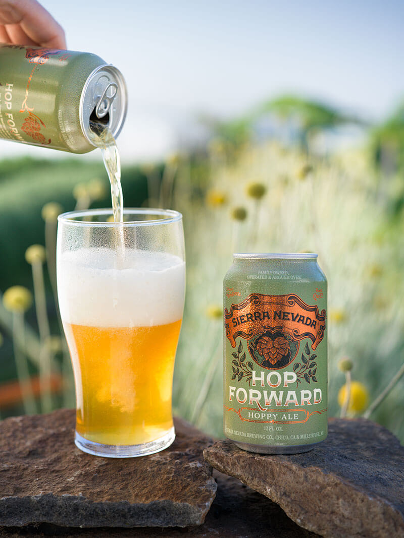 A hand pouring a can of Hop Forward into a glass, with garden flowers in the background