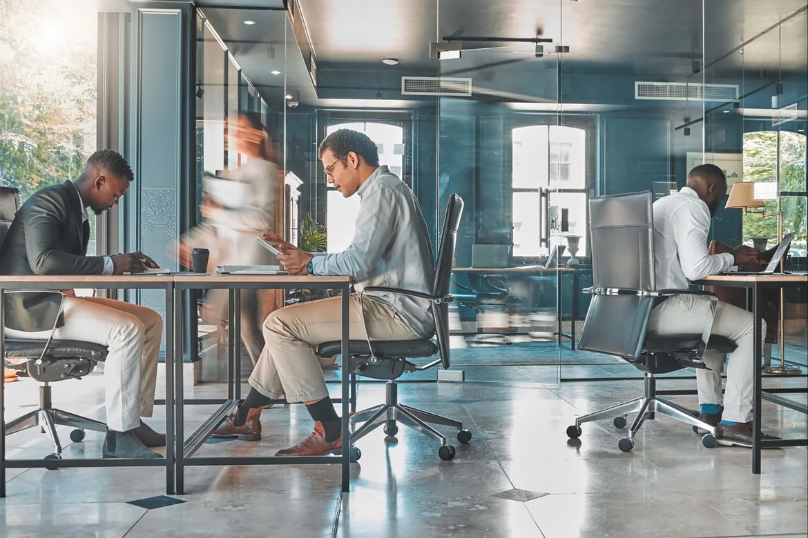 Modern office with three people working at desks in a glass-walled workspace; one person walks between tables, creating motion blur, while others focus on a tablet, paperwork and a laptop.